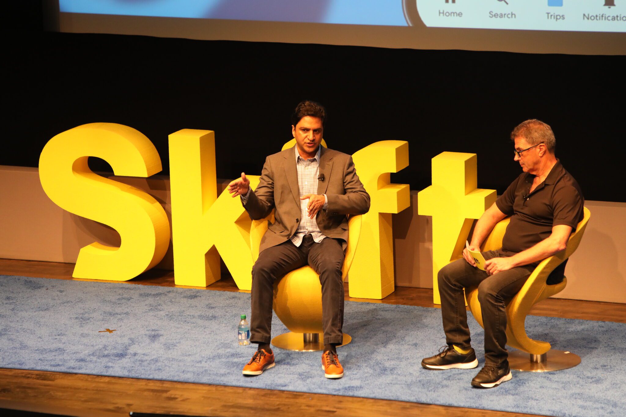 Two men on stage talking, sitting by yellow "Skift" sign